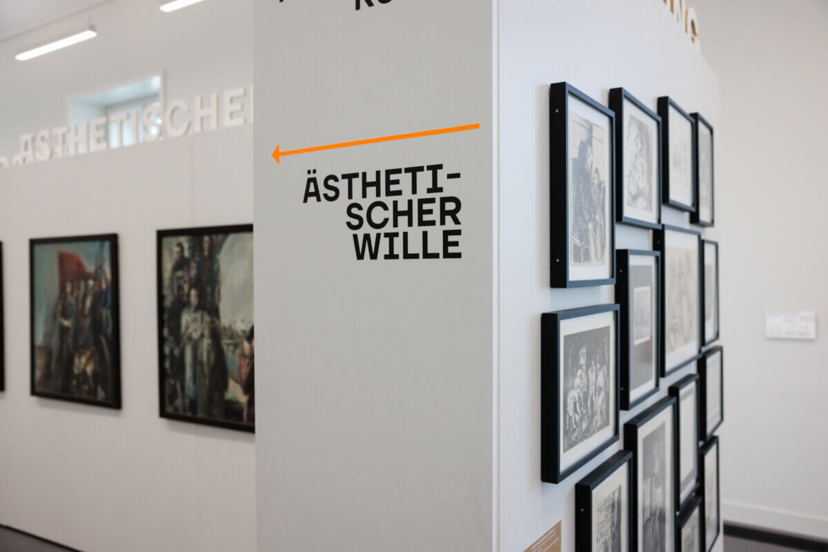 Ausstellungsansicht mit Bildwand auf der rechten Seite und zwei Gemälden auf der linken Seite. In der Mitte grenzt eine Säule/ein Wandteil mit der Aufschrift "Ästhetischer Wille" die beiden Zonen ab.