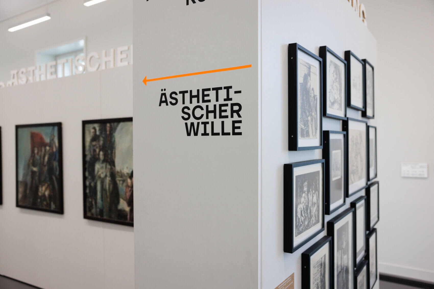 Ausstellungsansicht mit Bildwand auf der rechten Seite und zwei Gemälden auf der linken Seite. In der Mitte grenzt eine Säule/ein Wandteil mit der Aufschrift "Ästhetischer Wille" die beiden Zonen ab.