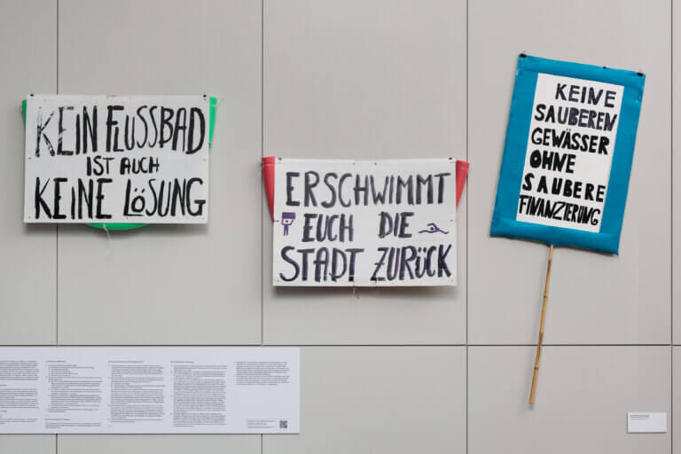Ausstellungsansicht mit drei Protestplakaten mit der Aufschrift "Kein Flussbad ist auch keine Lösung", "Verschwimmt euch die Stadt zurück" und "Keine sauberen Gewässer ohne saubere Finanzierung" an einer Wand. Daneben sieht man einen Ausschnitt des Gesetzentwurfs "Rechte der Spree".