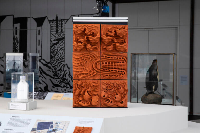 Ausstellungsansicht mit Keramik im Zentrum, außerdem Flasche mit MOF-Pulver links davon. Im Hintergrund ist ein Wandmotiv von Werner Heldt zu sehen