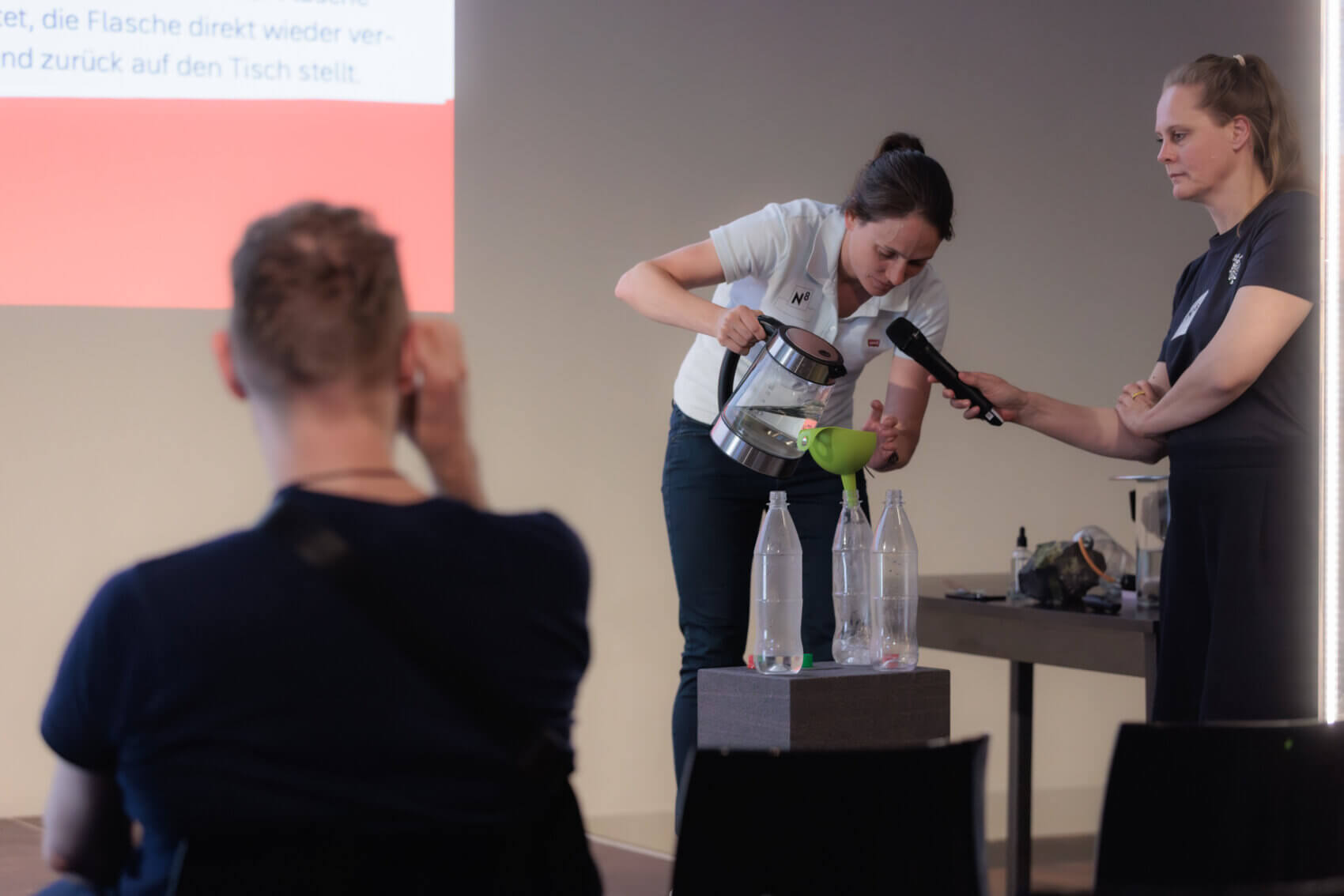 Eine Frau kippt mithilfe eines grünen Trichters Wasser aus einem Wasserkocher in eine leere Plastikflasche. Auf einem Podest vor ihr stehen noch zwei weitere Flaschen. Während sie das Wasser kippt, spricht sie in ein Mikrofon, das von einer weiteren Frau in schwarzem T-Shirt gehalten wird. Im Vordergrund ist ein Zuschauer und die Stuhlreihe erkennbar.
