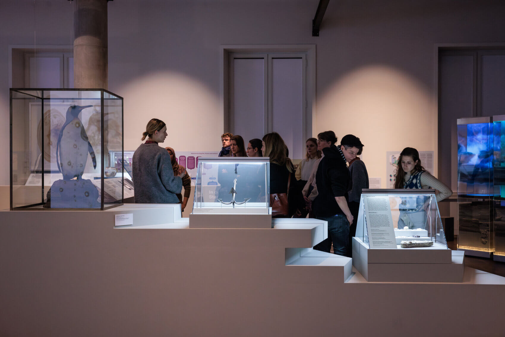 Eine Gruppe Menschen stehen auf einer Landschaft in der Ausstellung "On Water". Im Vordergrund sind verschiedene Objekte in Vitrinen zu sehen, darunter ein Pinguin, ein Anker und Steine.