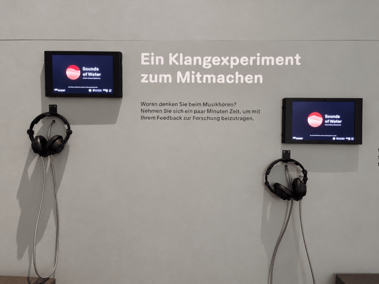 Zwei Bildschirme mit der Anwendung "Sounds of Water" hängen an einer grauen Wand in der Ausstellung. Neben Kopfhörern ist hier der Schriftzug "Ein Klangexperiment zum Mitmachen" zu sehen
