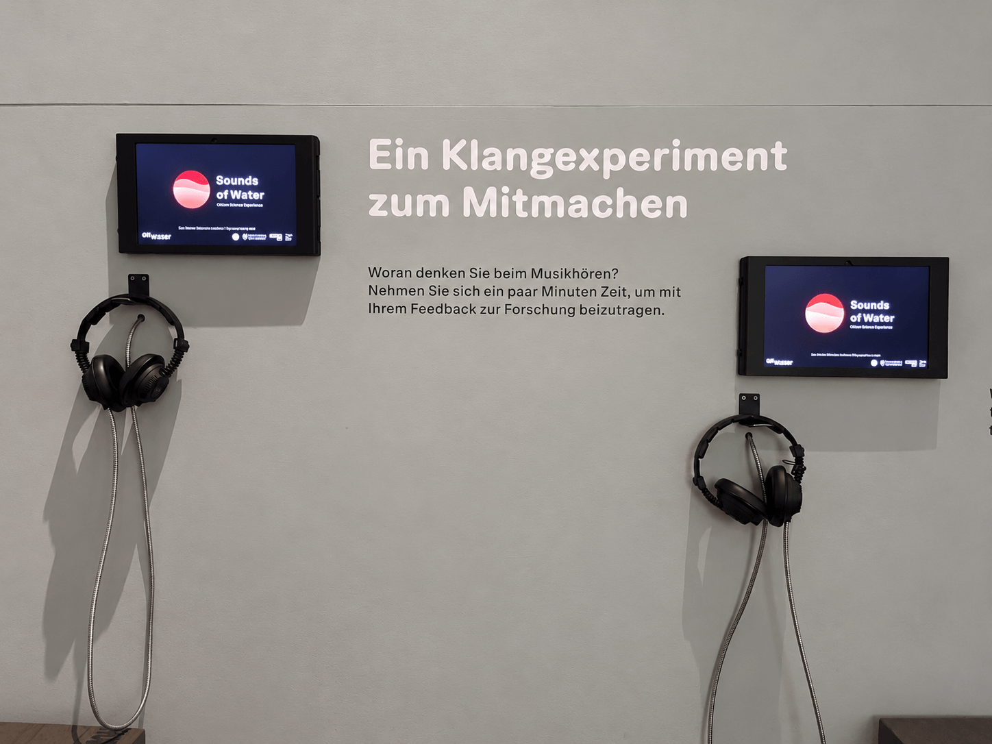 Zwei Bildschirme mit der Anwendung "Sounds of Water" hängen an einer grauen Wand in der Ausstellung. Neben Kopfhörern ist hier der Schriftzug "Ein Klangexperiment zum Mitmachen" zu sehen