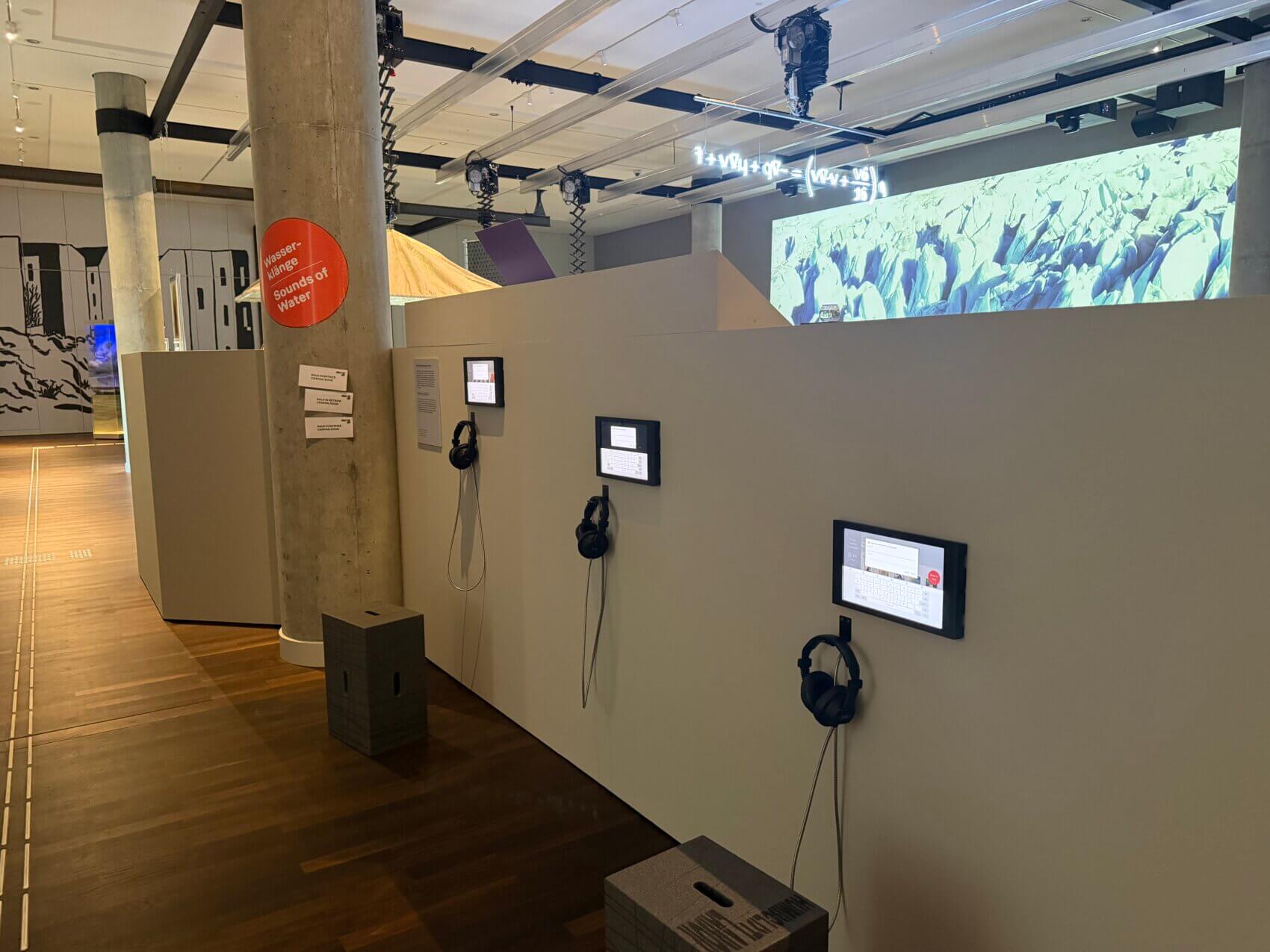 Ausstellungsansicht mit einer niedrigen Wand an der drei Bildschirme auf unterschiedlichen Höhen hängen. Neben den Bildschirmen hängen außerdem Kopfhörer. Vor ihnen stehen Hocker. Im Hintergrund ist die Medienwand mit einem Wassermotiv erkennbar.