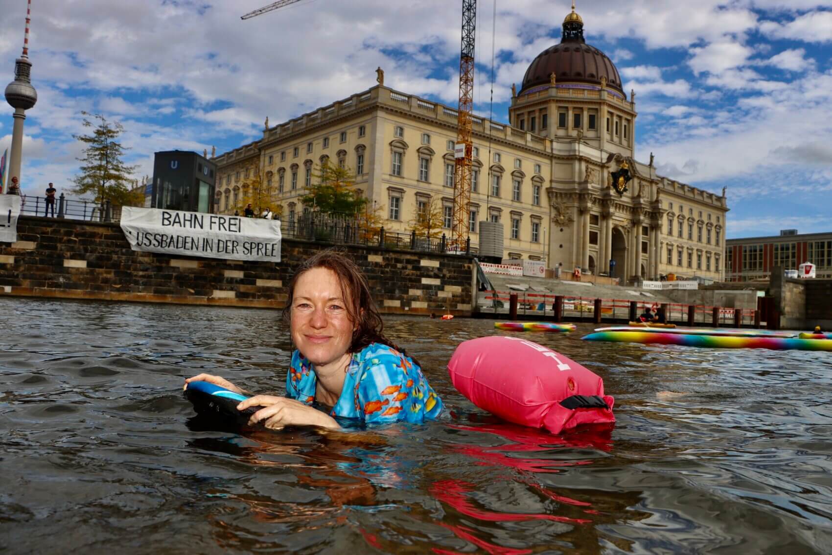 Eine Person mit blauem Oberteil auf denen Fische abgedruckt sind, schwimmt vor dem Humboldt Forum in der Spree.
